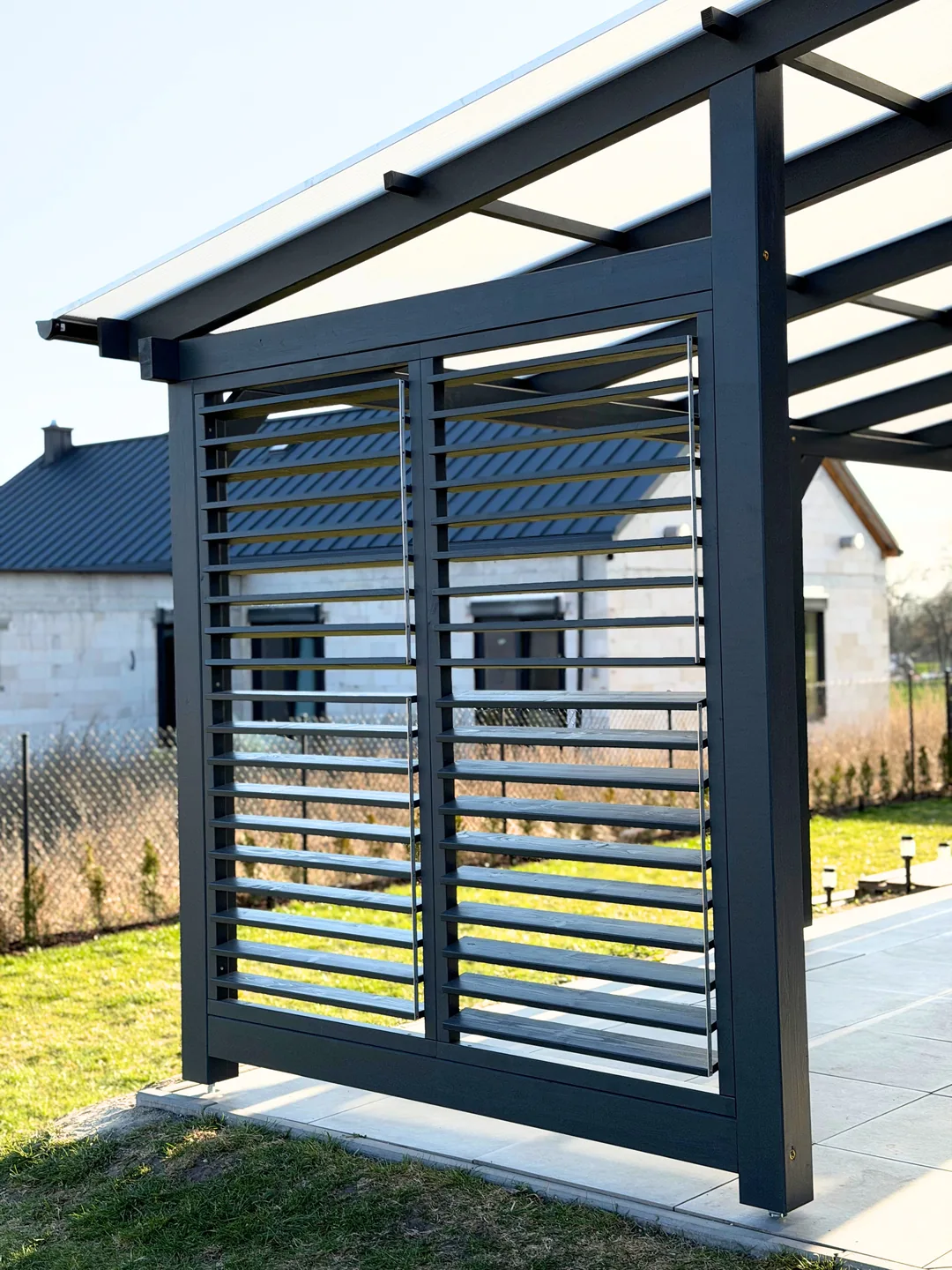 Pergola w kolorze antracytu z ażurową ścianką z lameli poziomych przy nowym domu