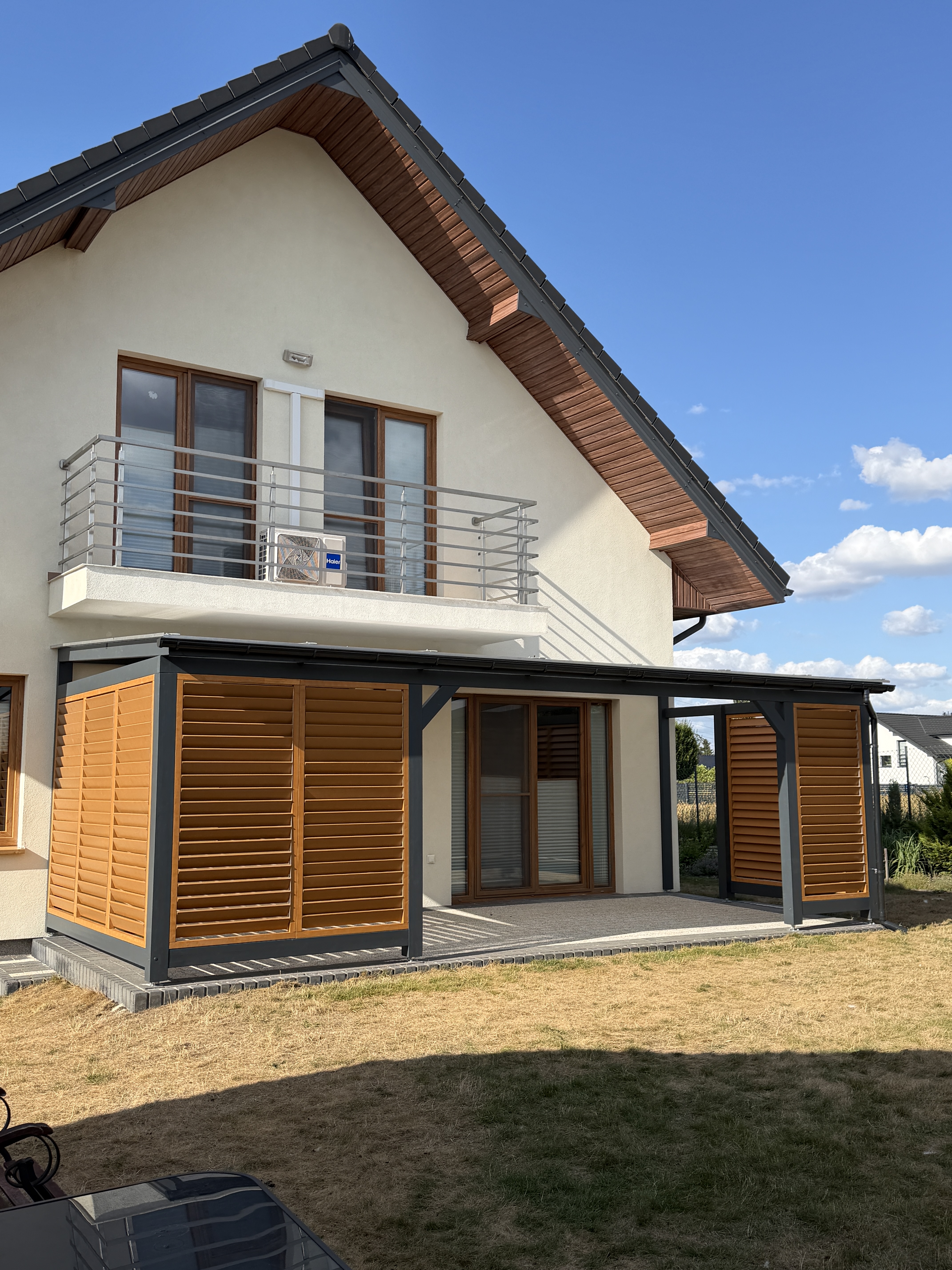Zadaszenie tarasu z antracytową konstrukcją i żaluzjami w kolorze dębu przy domu z balkonem